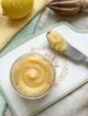 Lemon Curd Casero (Crema de Limón) Lemon curd casero servido en un frasco de vidrio con limones frescos y ralladura de limón sobre una superficie de mármol blanco.