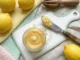 Lemon Curd Casero (Crema de Limón) Lemon curd casero servido en un frasco de vidrio con limones frescos y ralladura de limón sobre una superficie de mármol blanco.
